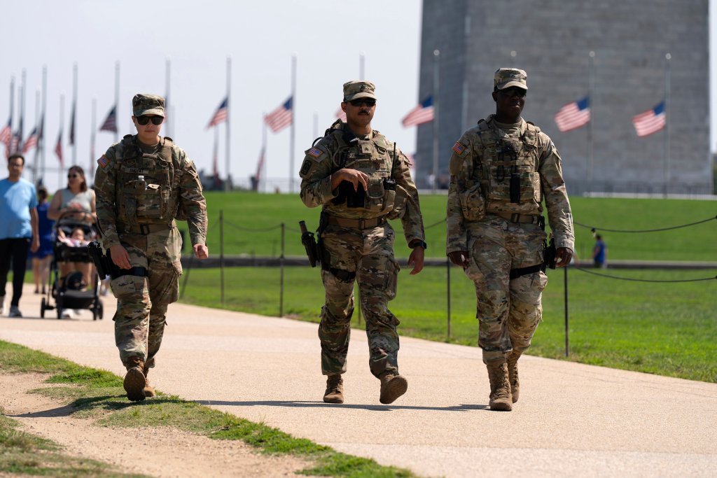 Three people in uniform walk along a sidewalk. Behind them a row of half-staff American flags wave in the breeze.