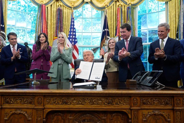 People clap surrounding a man sitting at a large desk holding up two pieces of paper.