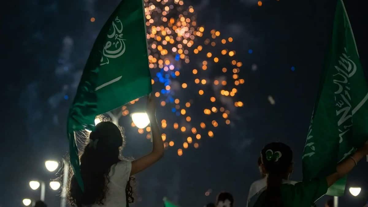 Silhouettes of children waving Saudi flags against a night sky filled with colorful fireworks, celebrating the 95th National Day, with streetlights illuminating the scene below.
