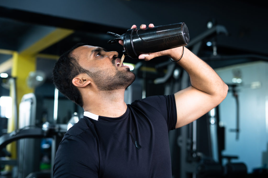 bodybuilder drinking or taking protein powder and mixing with water on bottle by shaking at gym - concpet of muscular gain...