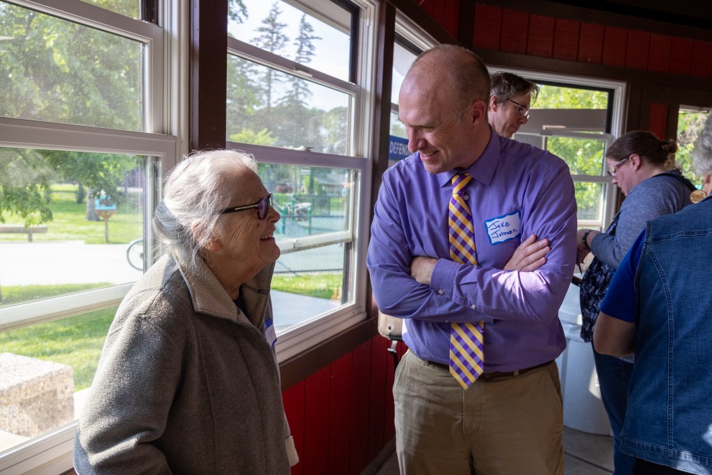 a main in a blue shirt and yellow ties has his arms folded as he speaks with a woman