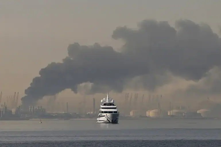 Thick black smoke rises from Dubai’s Jebel Ali port as a yacht sails nearby.