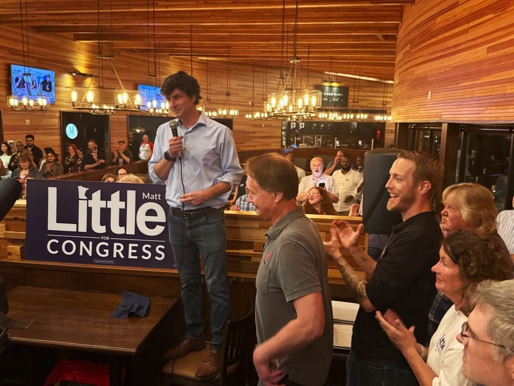 man speaking into a microphone near a matt little for congress sign as a crowd applauds