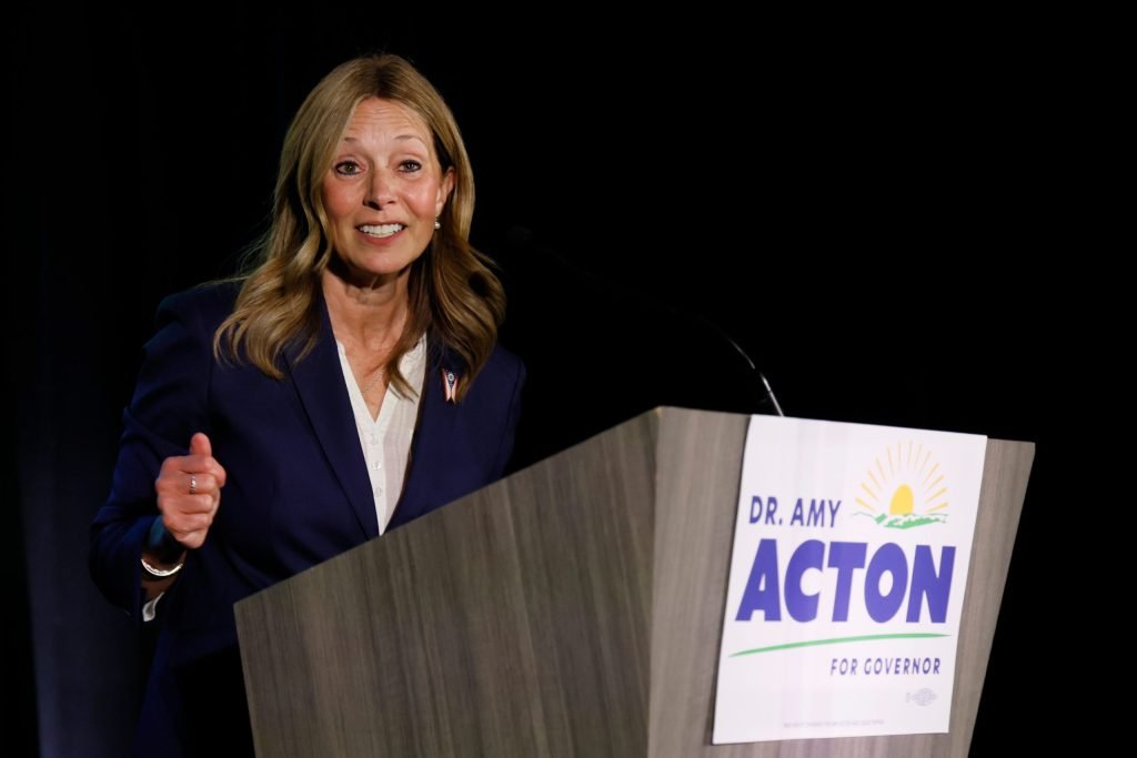 woman speaking at a podium with a dr amy acton for governor sign attached to the front