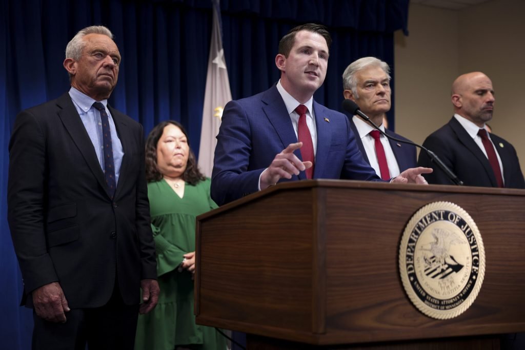 man speaking at a department of justice podium during a press conference with officials standing behind him