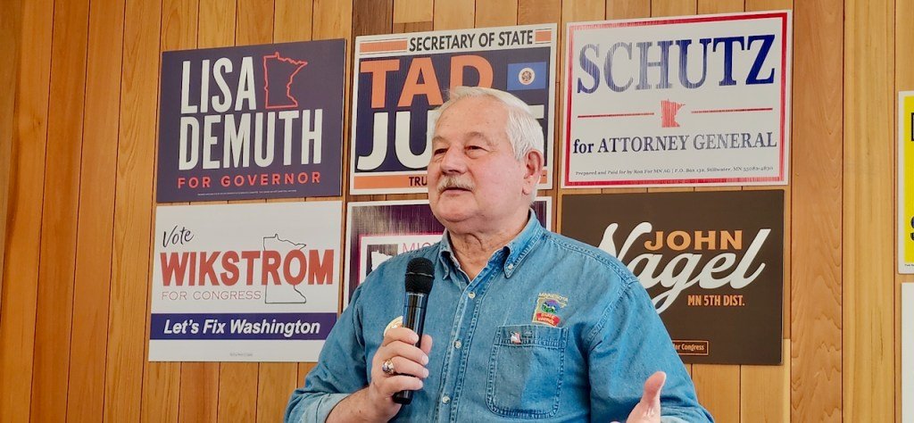 john nagel holding a microphone speaking in front of political campaign signs on a wood paneled wall